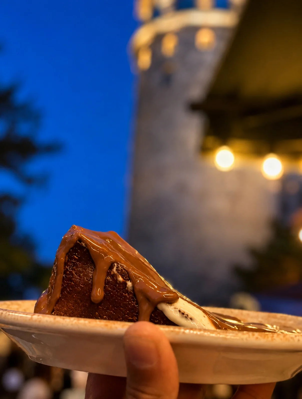 A hand holding a spoon over a plated dessert on a terrace table at blue hour
