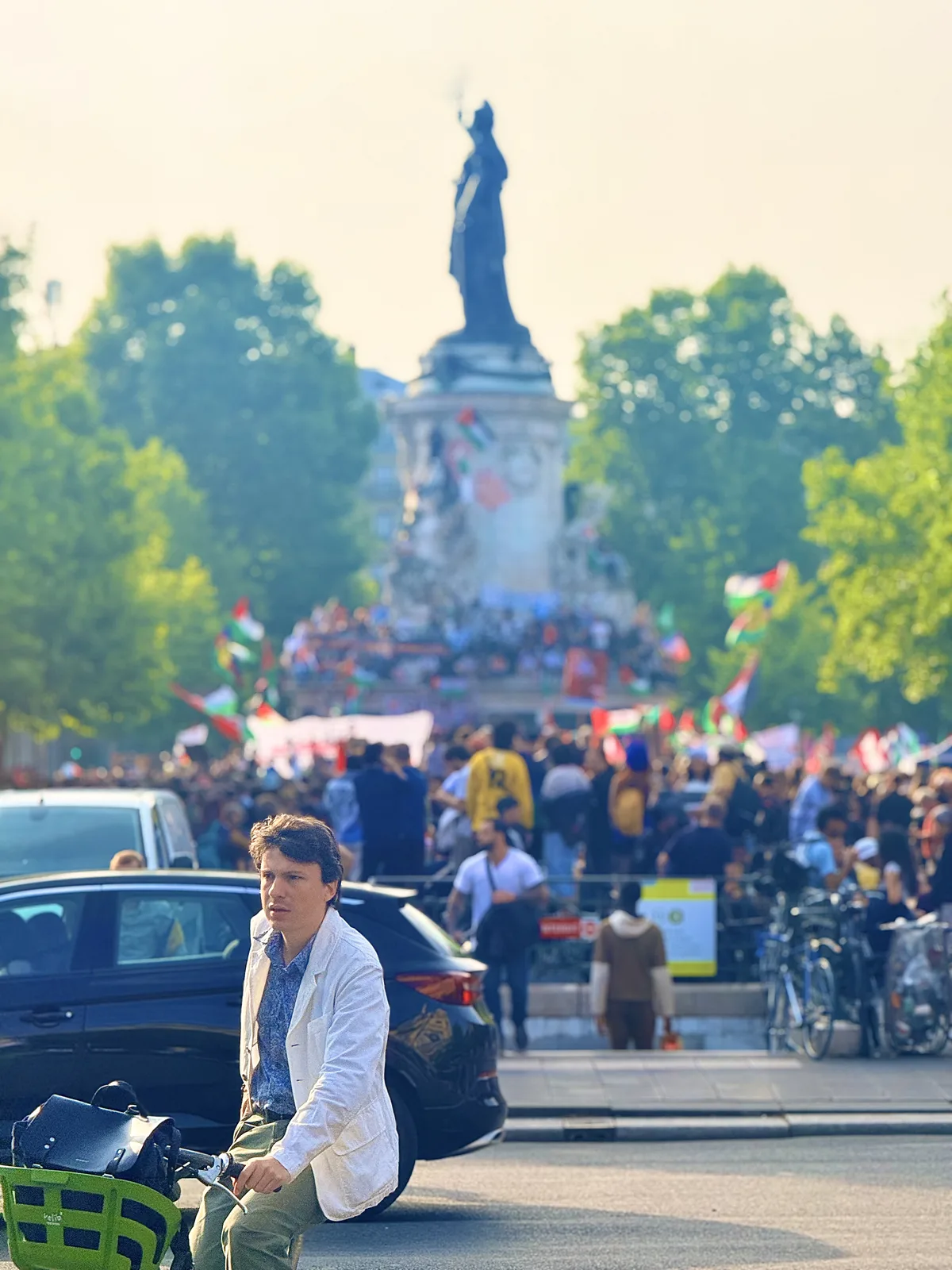 A cyclist pausing on a Paris street as a flag-filled crowd gathers around the Place de la République monument